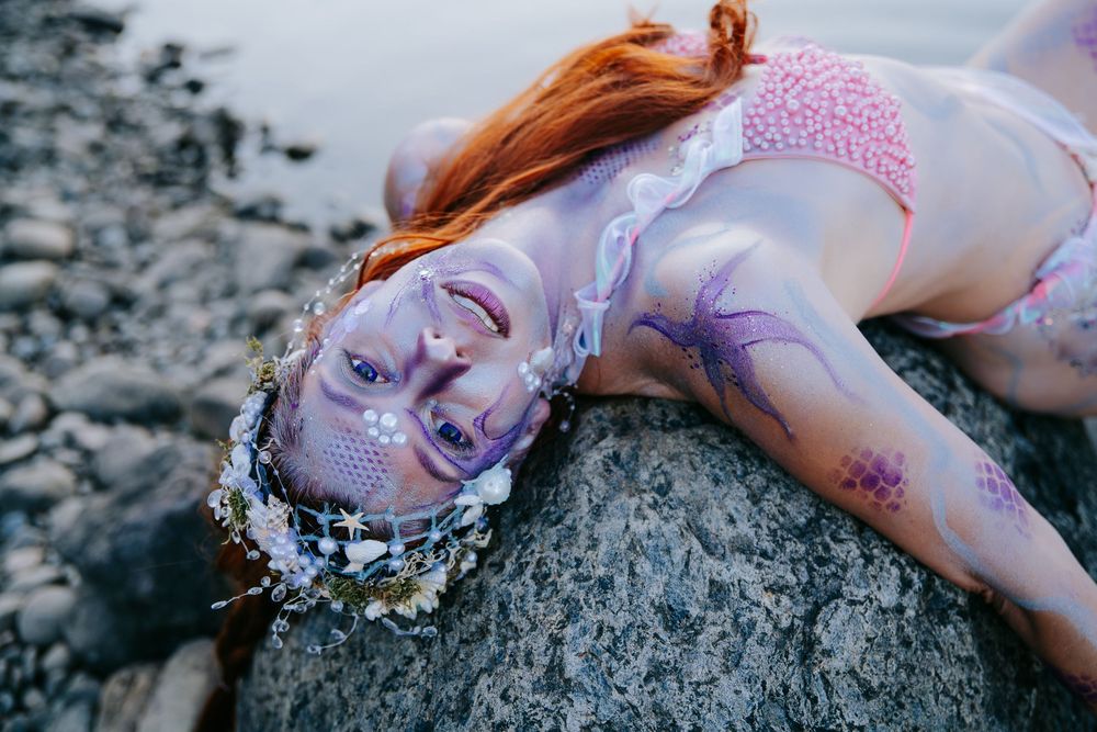 A person with red hair lies on dark rocks near water in an artistic fashion photography series.