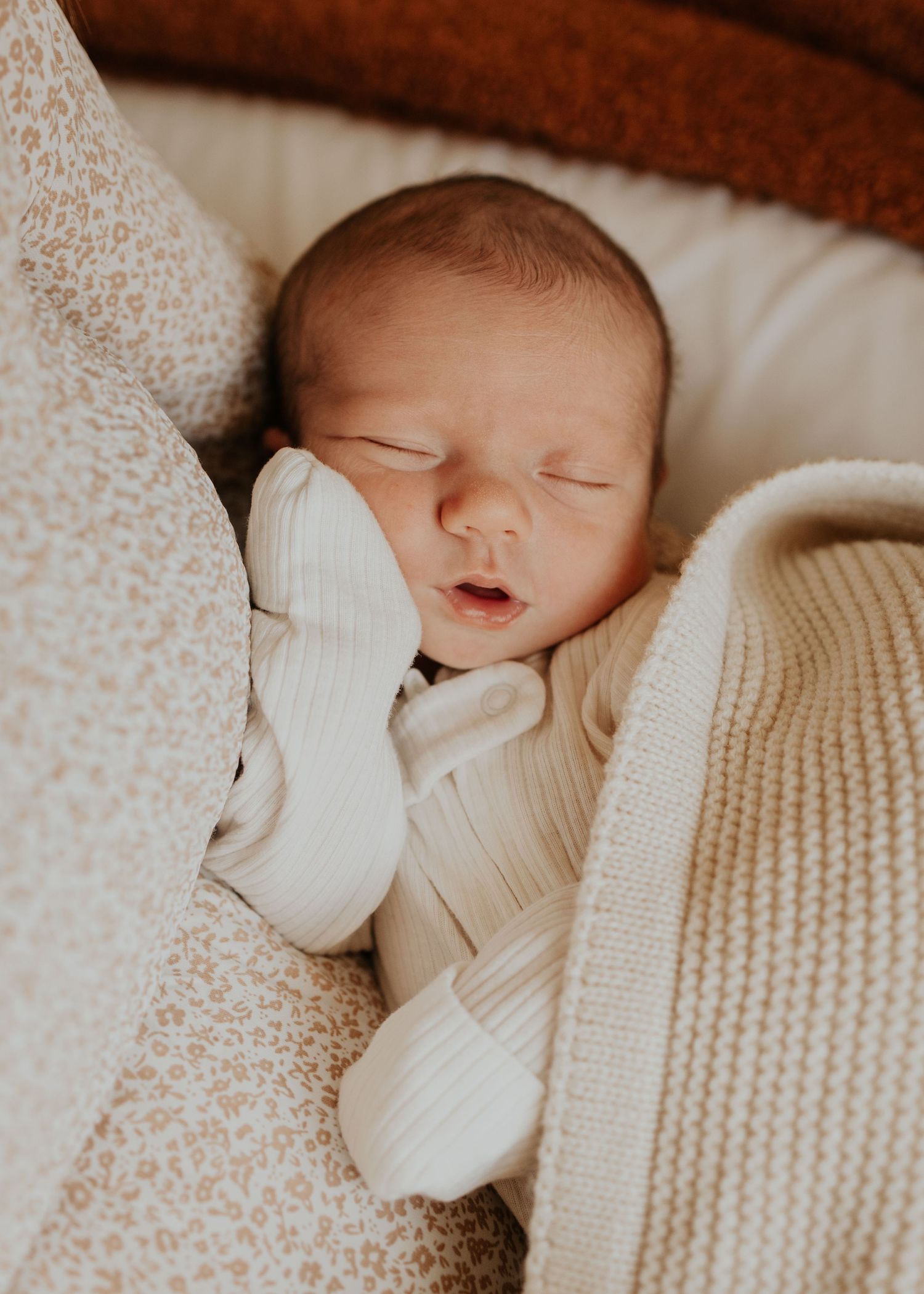 A sleeping newborn baby wrapped in cozy white knit blankets on a textured cream-colored surface during newborn photography session.