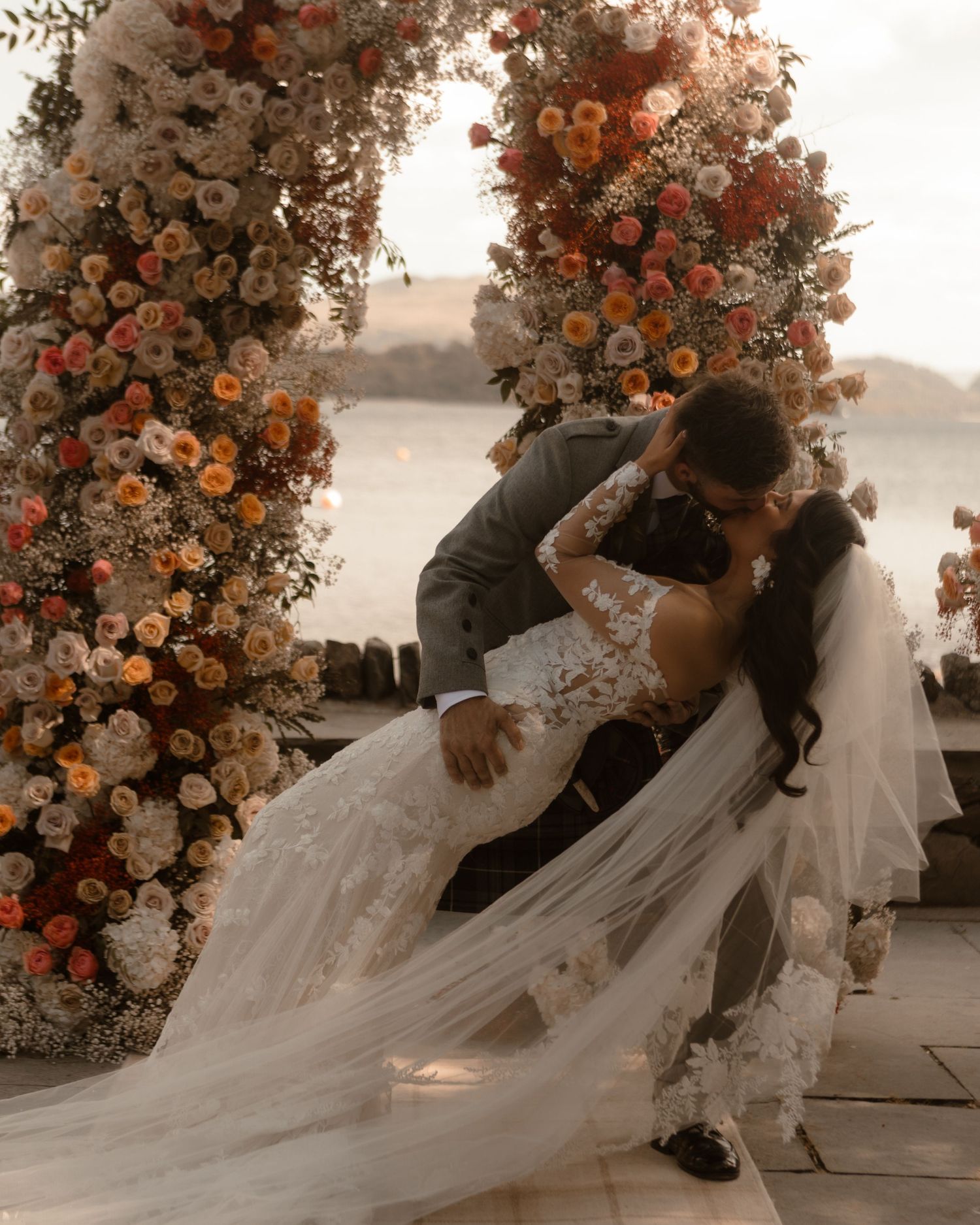 A dramatic wedding kiss under a beautiful floral arch with orange and pink flowers at sunset.