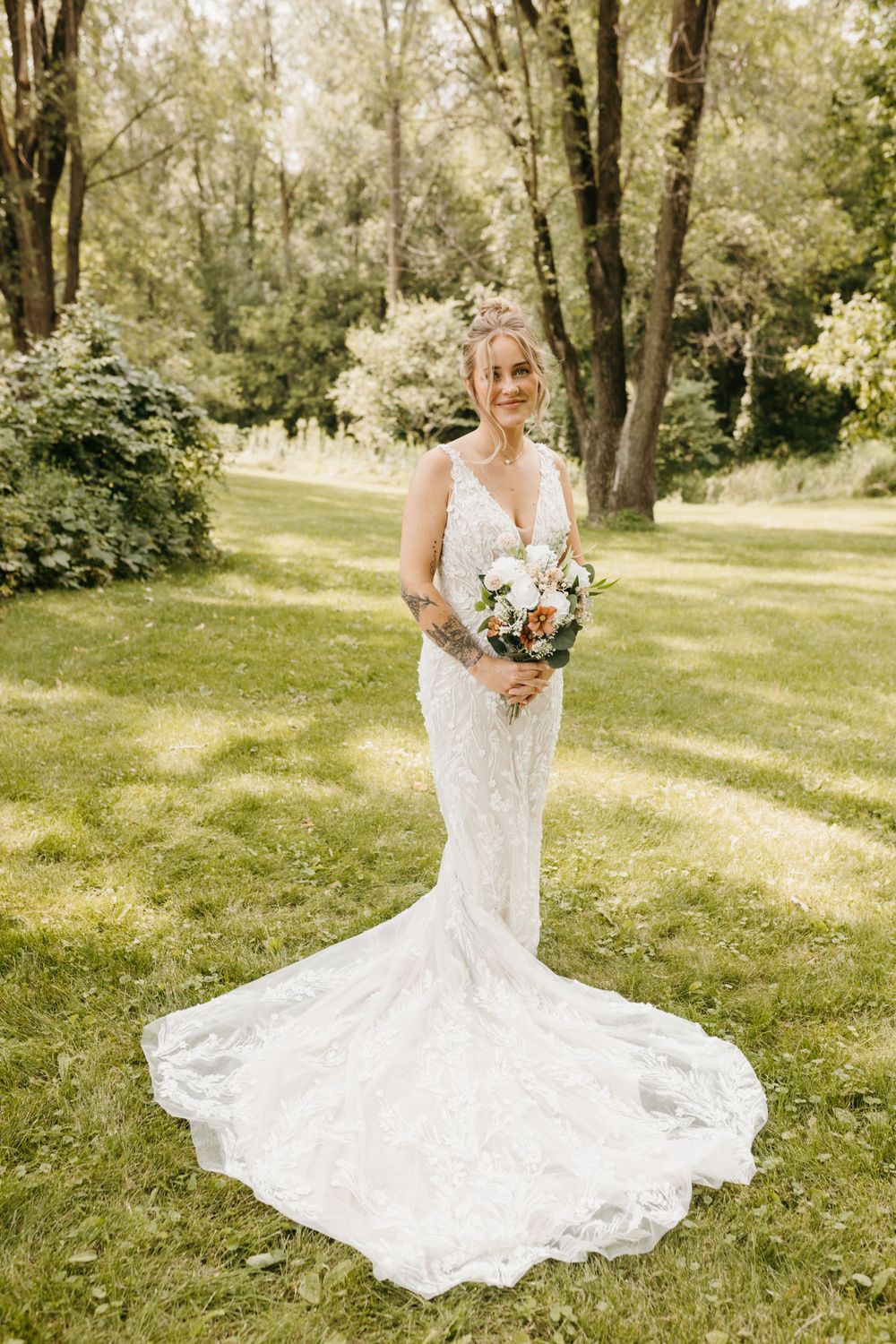Smiling bride in a lace fit-and-flare wedding gown holding a bouquet in a garden.