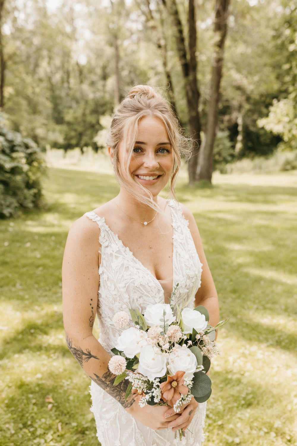 Radiant tattooed bride smiling in a lace V-neck wedding dress holding a white rose bouquet.