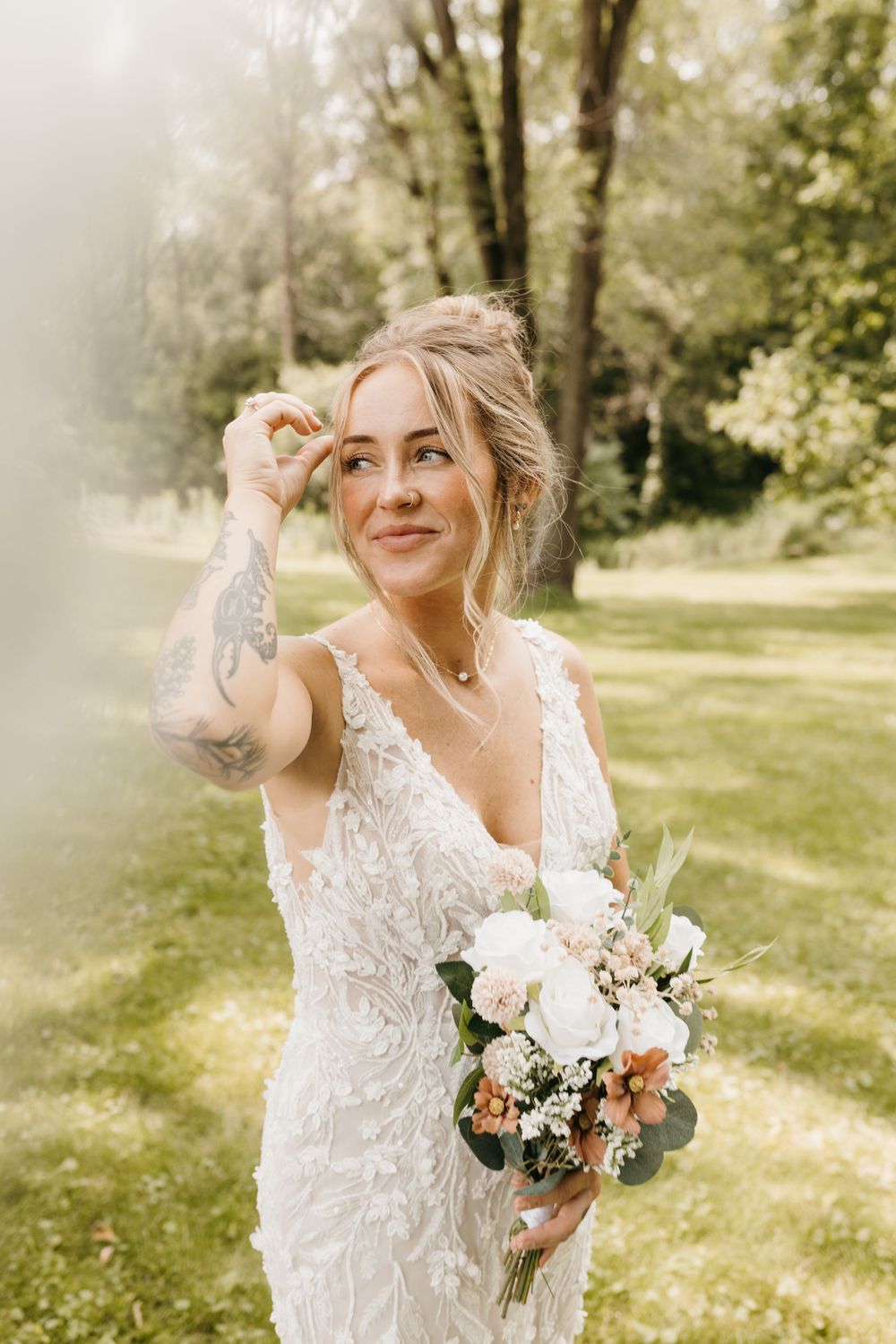 Tattooed bride in a lace wedding gown gazing upward while holding a bouquet outdoors.
