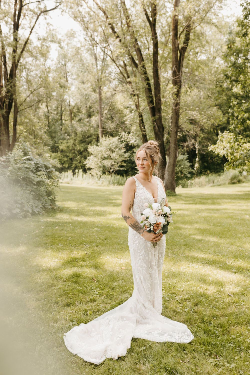 Tattooed bride in a lace wedding dress with a long train standing in a lush garden.