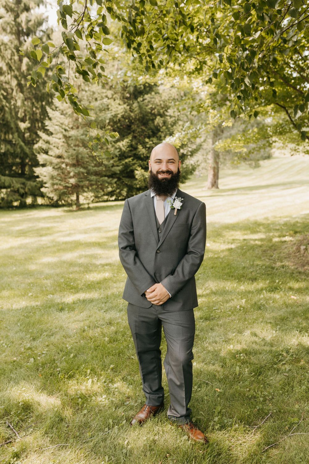 Bald bearded groom in a charcoal suit with boutonniere smiling outdoors on green lawn.