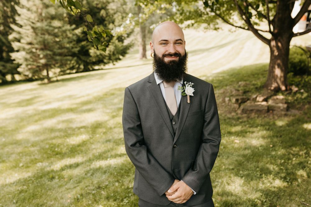 Smiling bald groom with full beard wearing gray suit and white boutonniere outdoors.