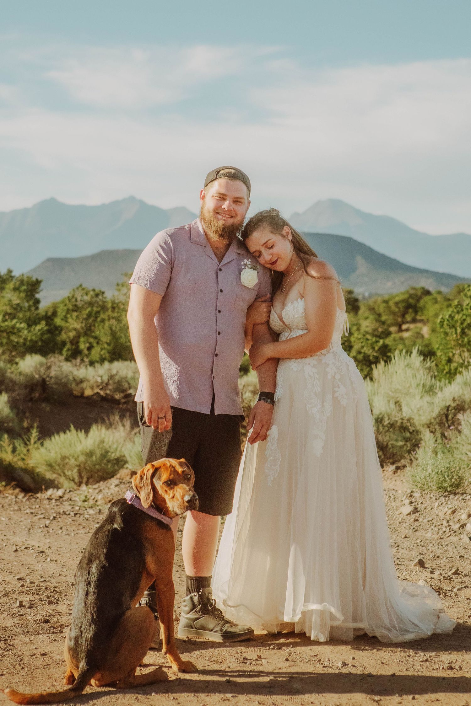 Bride in a flowing gown leans on her groom in a scenic mountain landscape, their dog sitting beside them.