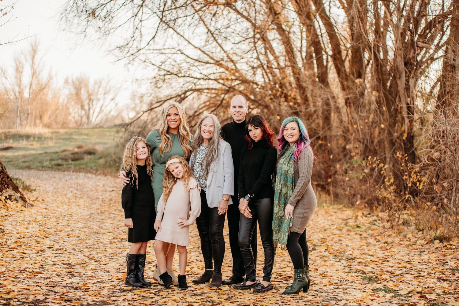Extended family portrait outdoors with bare trees and fallen leaves in autumn setting.