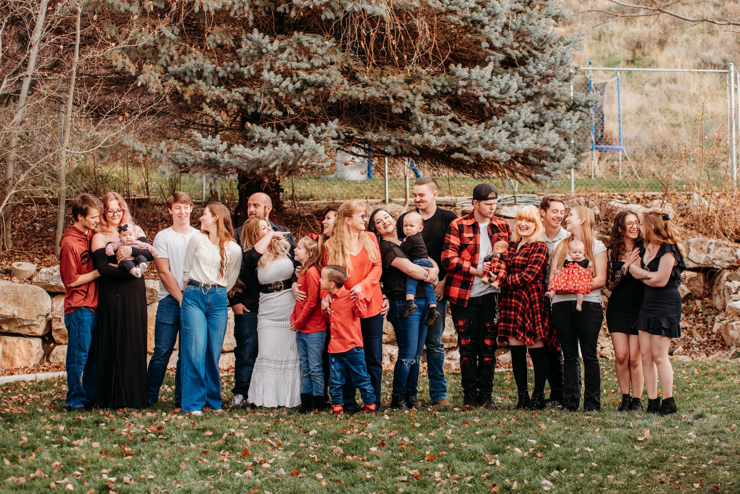 A large group of people in casual attire gathered outdoors for a photo near evergreen trees.
