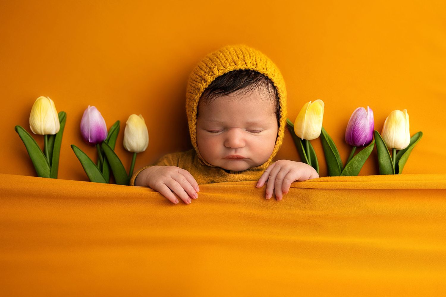 Sleeping newborn wearing yellow knit hat nestled in orange fabric with colorful tulips arranged alongside.