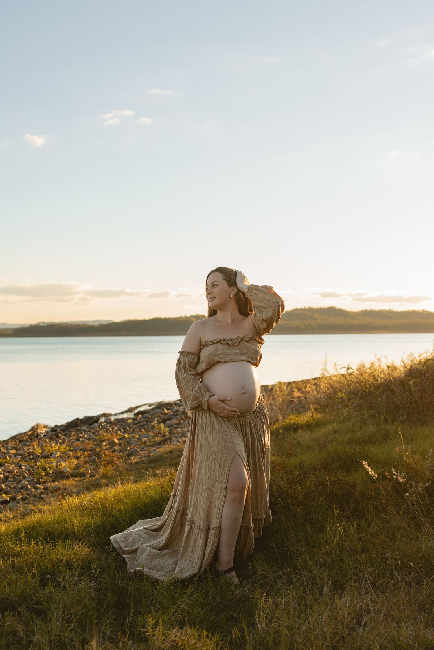 Maternity photoshoot at sunset shows expectant mother in flowing beige dress along grassy coastal shoreline.