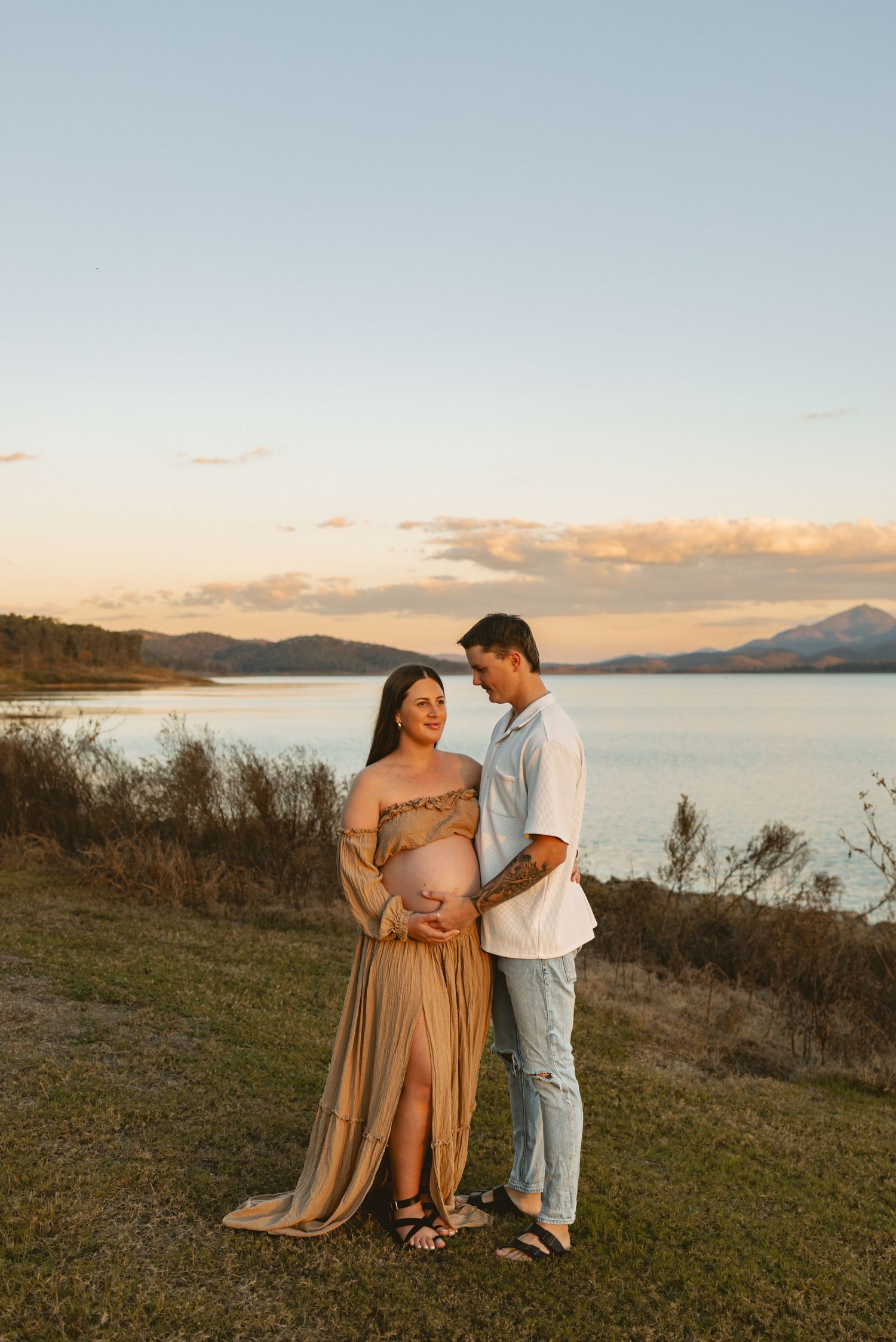 A couple shares a tender sunset moment on a grassy hillside overlooking a serene lake with mountains in the distance.