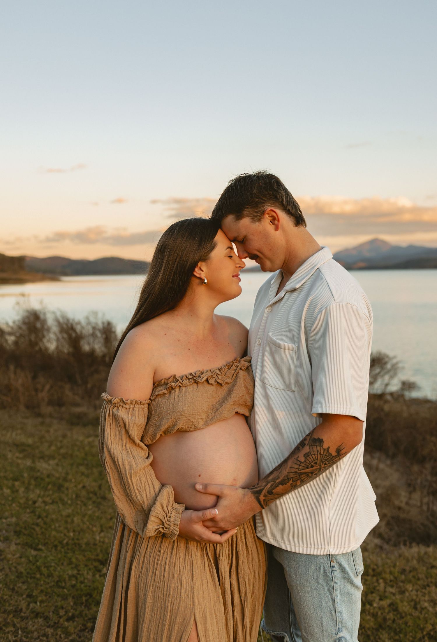 Couple shares intimate moment at sunset on grassy hillside overlooking ocean landscape in warm earth tones.