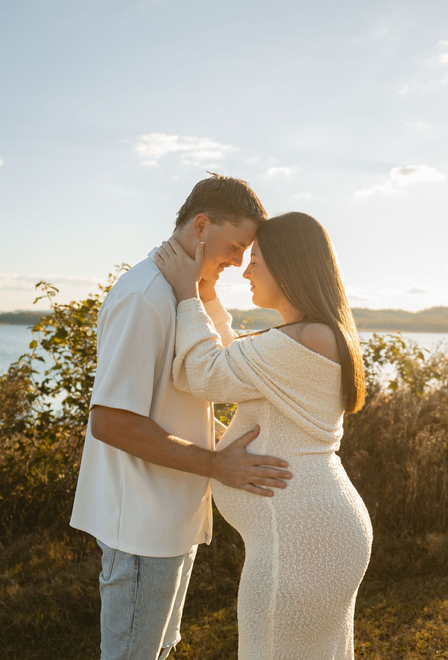 A couple shares an intimate outdoor maternity photoshoot at sunset, highlighting the mother's pregnant belly.