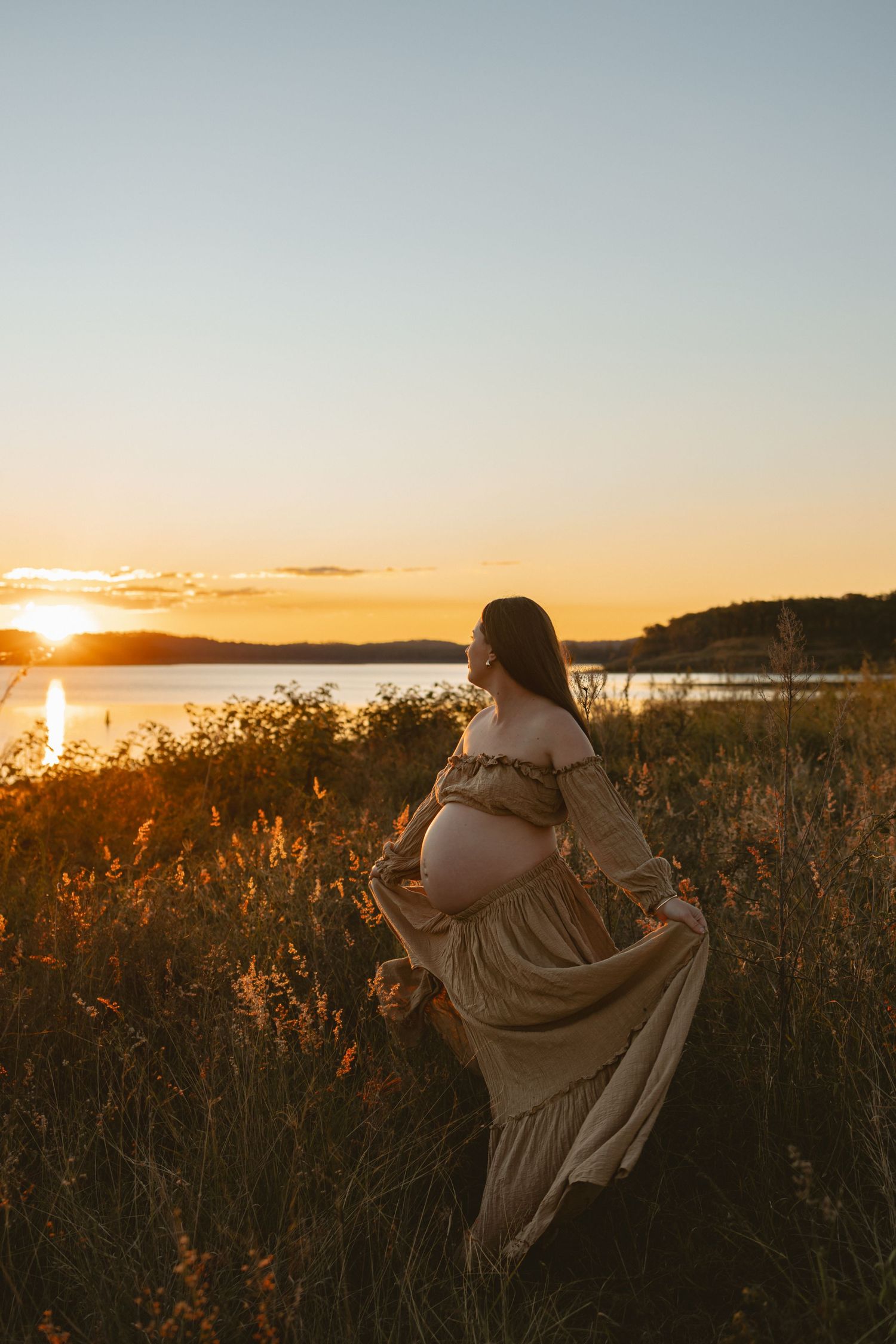A silhouette sits in tall grass on a hill overlooking a serene sunset over the water.