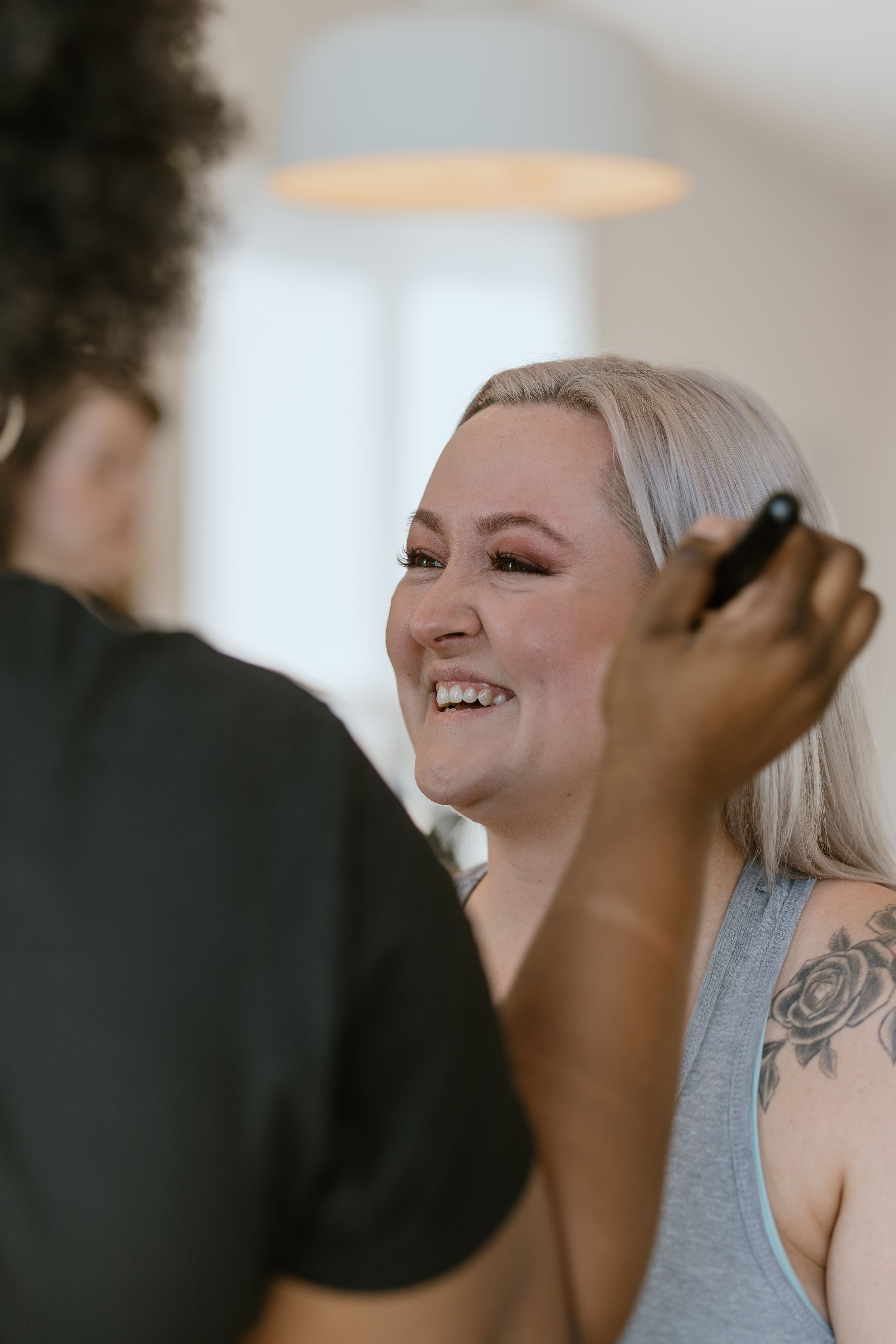 A person with platinum blonde hair and tattoos smiles joyfully while getting their makeup done by an artist.