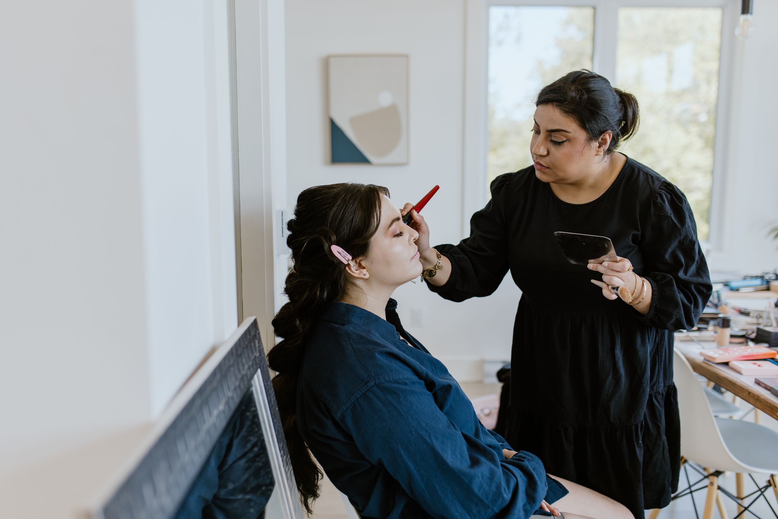 A makeup artist in black applies cosmetics to a client seated in a chair by a window for a wedding.