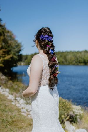 Beautiful white lace wedding dress photographed from behind with colorful flowers woven into long braided hair by a lake.