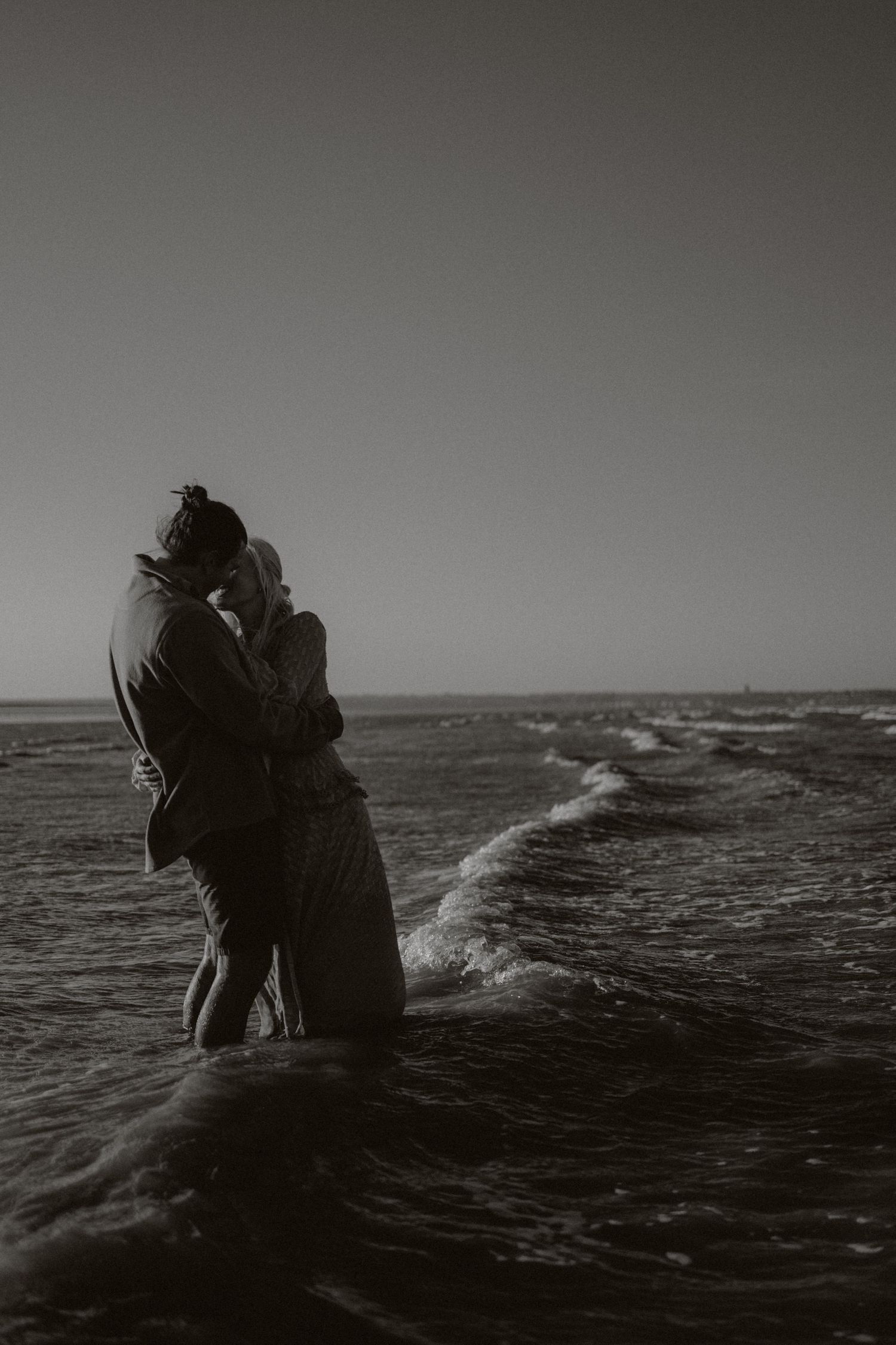 Silhouetted couple embracing against dramatic ocean waves in black and white.