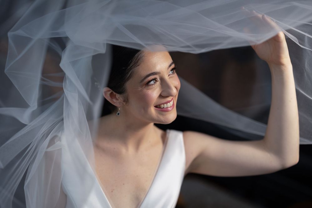 Smiling bride in v-neck wedding dress lifts her white veil gracefully in a candid moment.