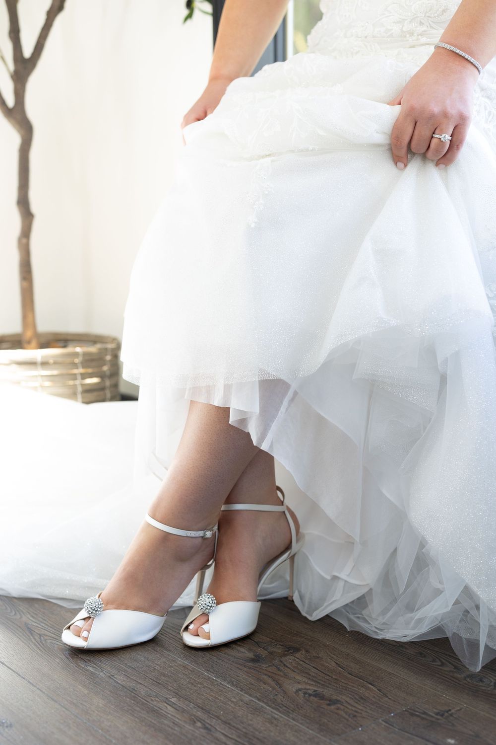 Close-up shots of elegant white bridal shoes with flowing tulle dress hem on dark hardwood flooring.