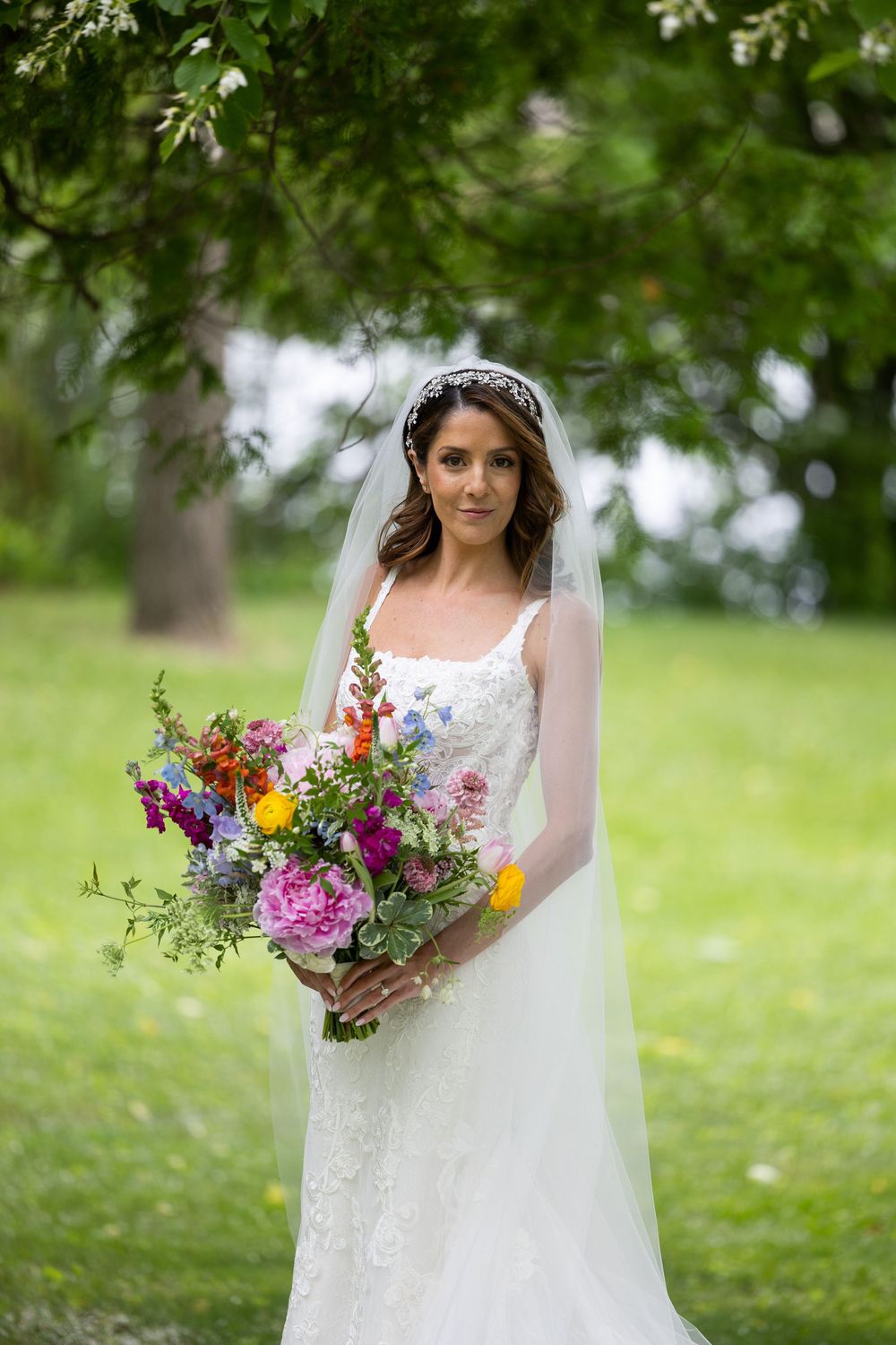 Beautiful bride in white lace dress holding vibrant wildflower bouquet on outdoor lawn with tree bokeh background.