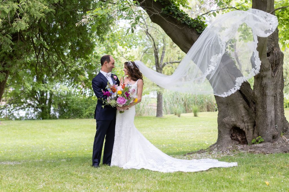 A couple in wedding attire embrace under a flowering tree while a white veil flows in the breeze.
