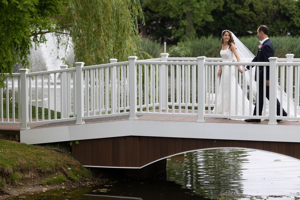 Two people in wedding attire stand on a white wooden bridge over water with a weeping willow tree in the background.