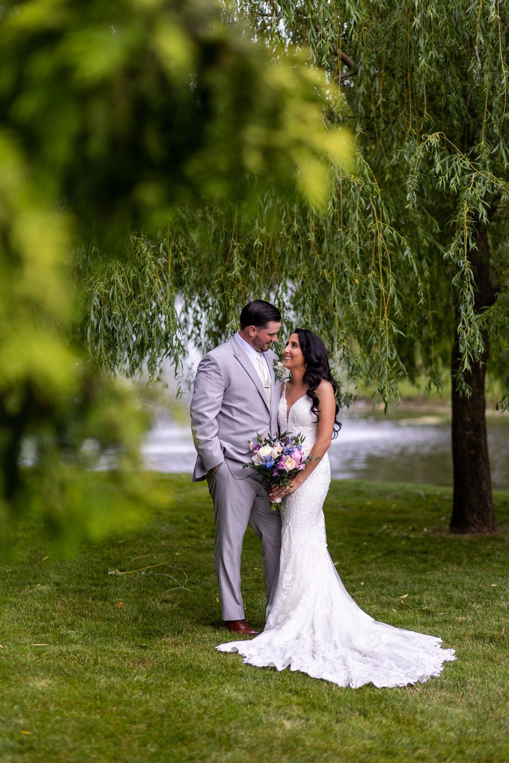 A romantic wedding couple stands beneath a weeping willow tree with a beautiful bouquet in a serene outdoor setting.