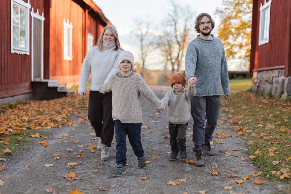Family of four walking hand-in-hand on an autumn path beside red Scandinavian buildings with fallen leaves.