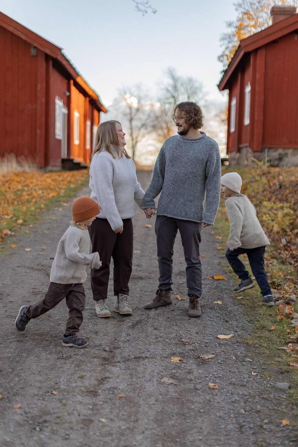 Family of four holding hands on a gravel path between red wooden buildings during autumn sunset.