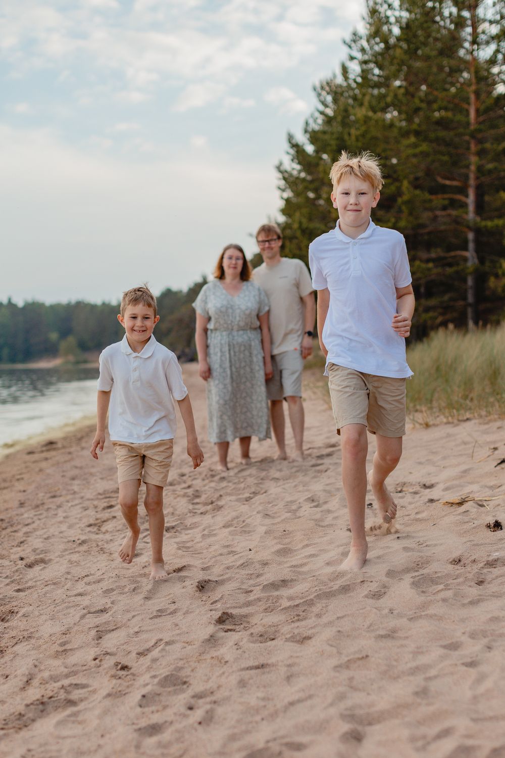 Two boys running barefoot on sandy beach with parents walking behind near pine trees.