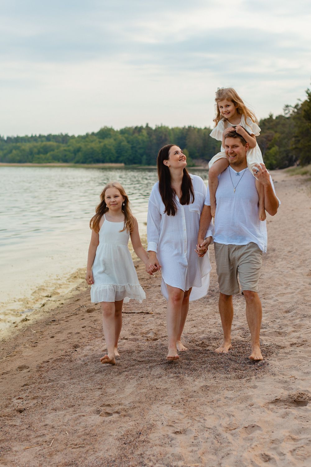 Family of four walking on sandy beach at sunset, father carrying daughter on shoulders. Perhekuvaus rannalla.