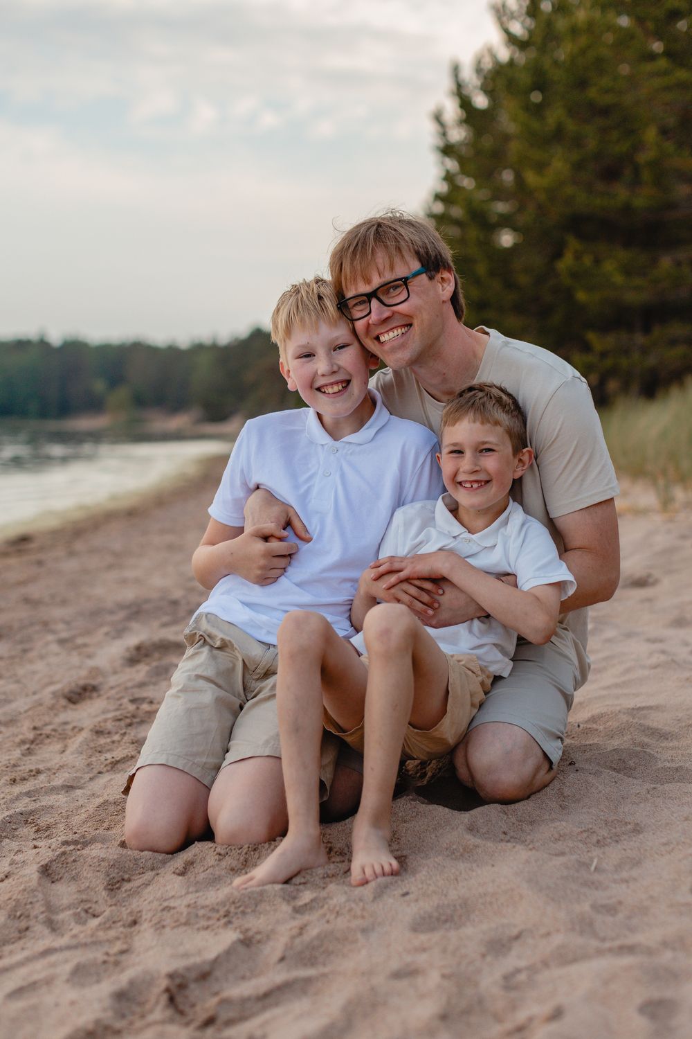 Father sitting on sandy beach hugging his two young blonde sons, all smiling and wearing white shirts.