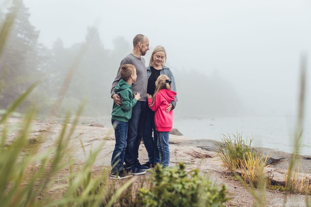 Family of four standing together on rocky shore in misty foggy weather near water.