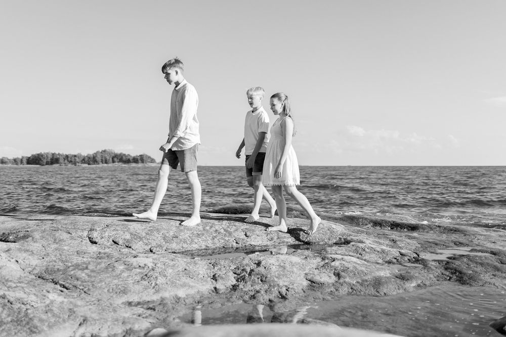 Perhekuvaus rantakallioilla. Perheen nuoriso kävelee laakeilla rantakallioilla, mustavalkoinen otos. Valokuvaaja Annina Nygård, Uusimaa.
