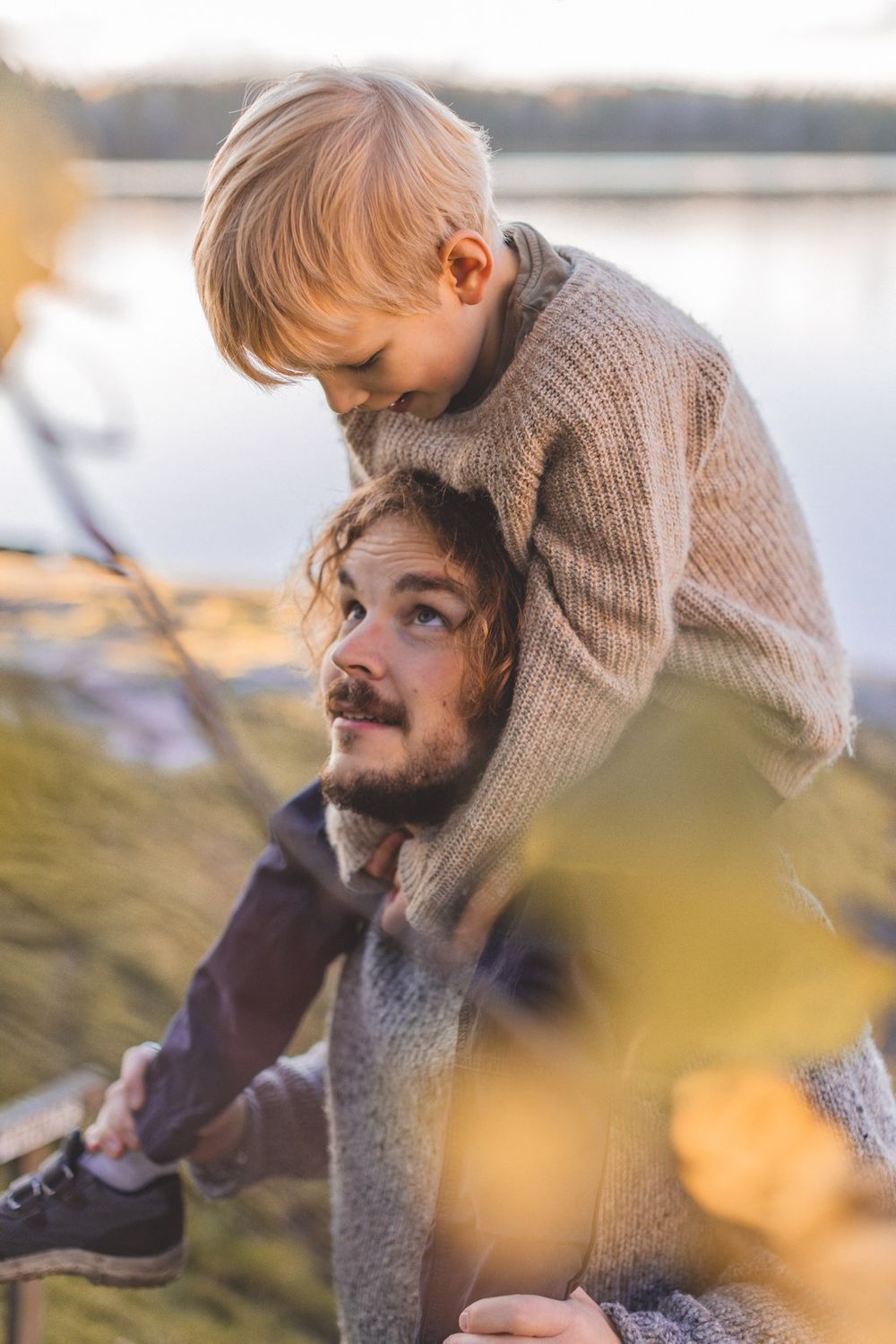 A bearded father carries his young blonde son on his shoulders near a serene lake during golden hour.