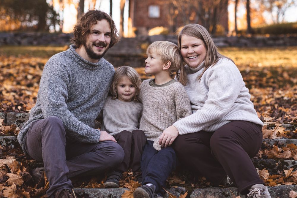 Family of four smiling outdoors in autumn, sitting among fallen leaves in coordinating neutral sweaters.