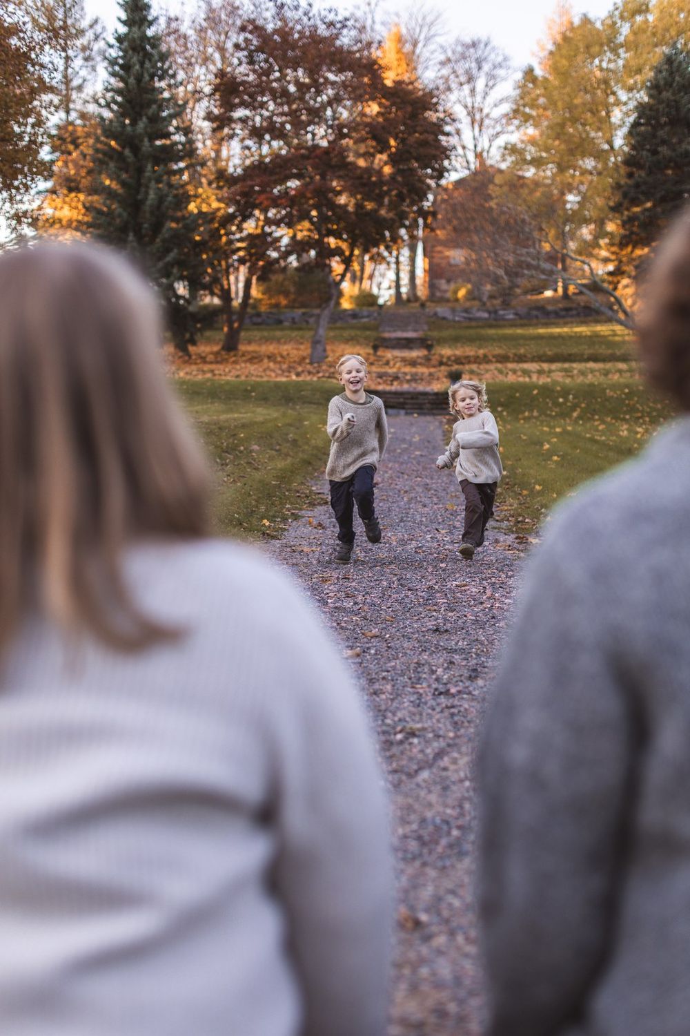 Two young children running joyfully toward adults on a gravel path in a colorful autumn park.
