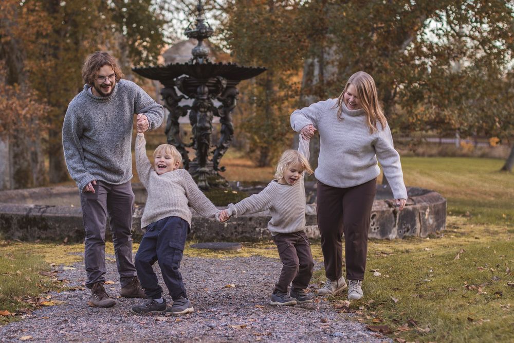 Family of four holding hands and playing on an autumn park path with a decorative fountain in the background.