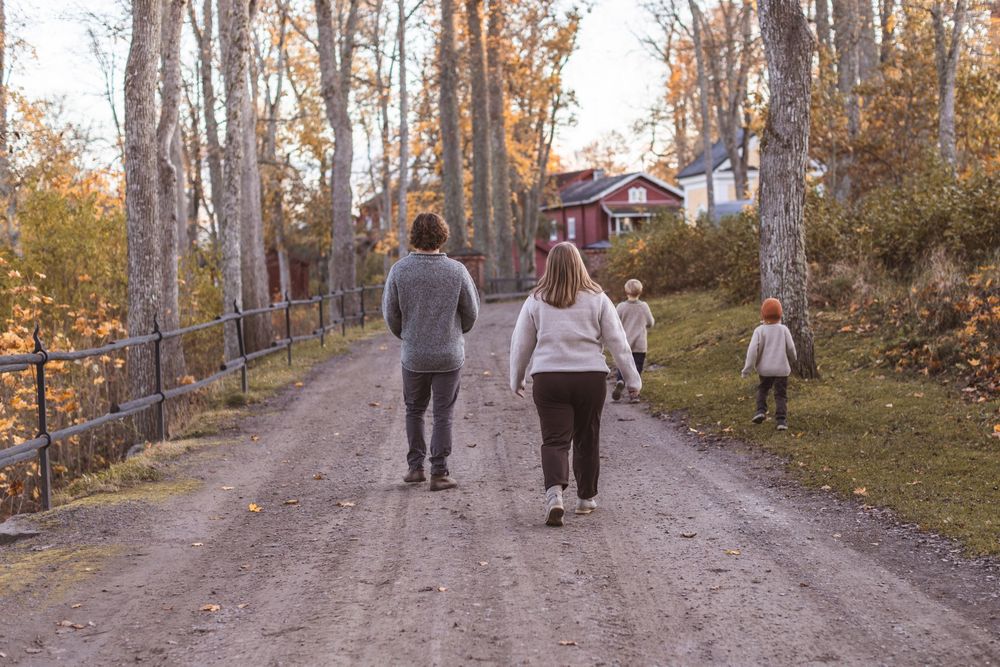 Family walking down a rustic dirt road surrounded by autumn trees toward a red Scandinavian house.