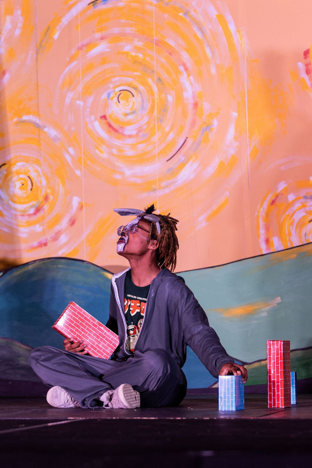 A performer lies on stage against an orange backdrop with spiral patterns.
