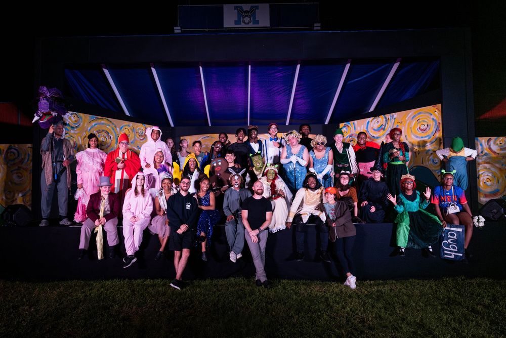 Large group photo of performers in colorful outfits on stage with dramatic lighting.