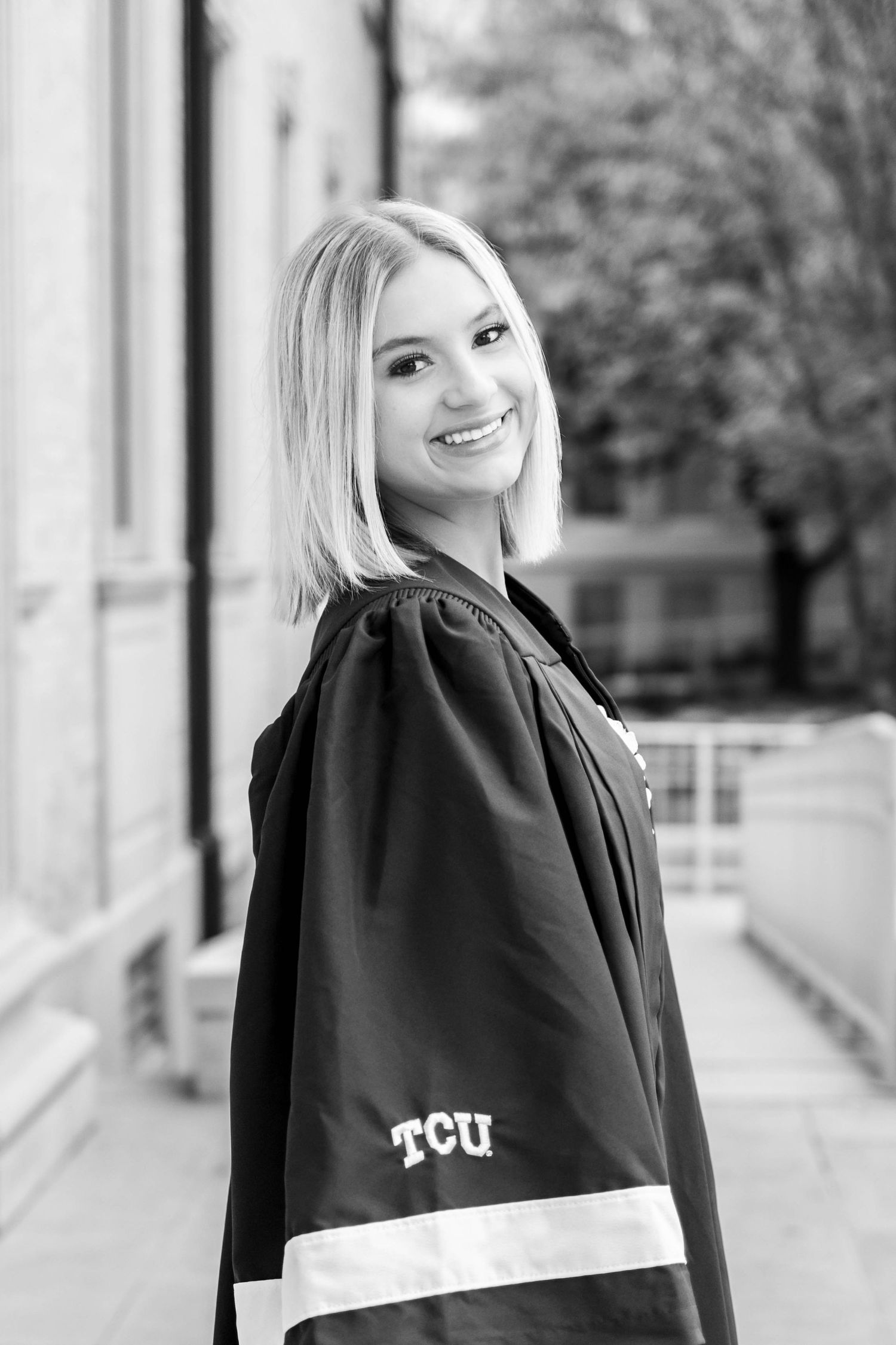 A graduate wearing a TCU academic gown smiles in a black and white portrait on a university campus.
