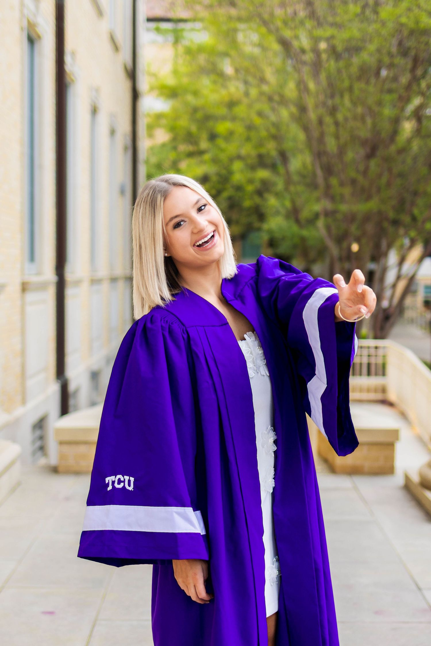 Graduate in purple TCU academic regalia smiles and gestures while standing outside a building.