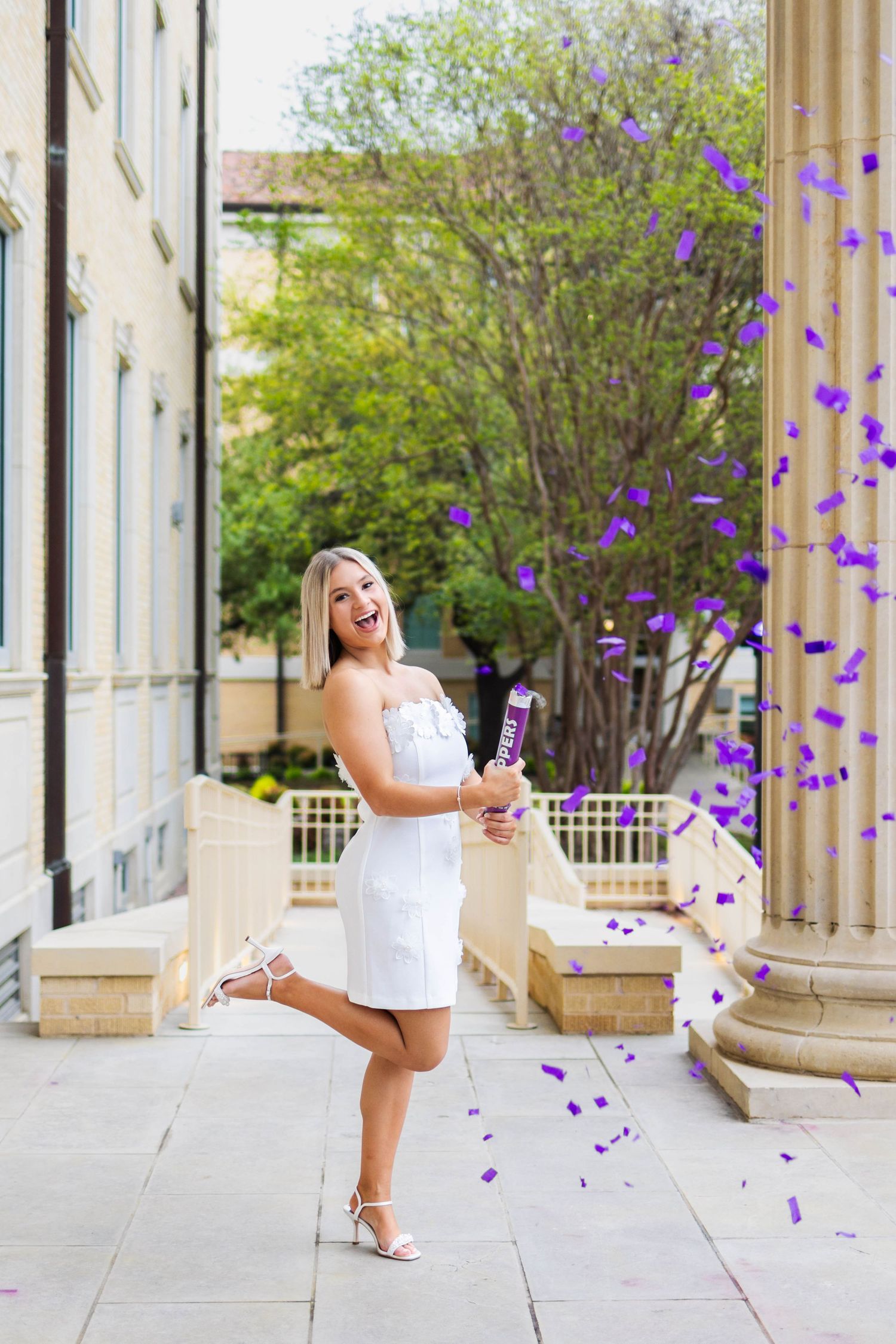 Person in white dress celebrates joyfully with purple confetti flying through the air near stone columns on a sunny day.