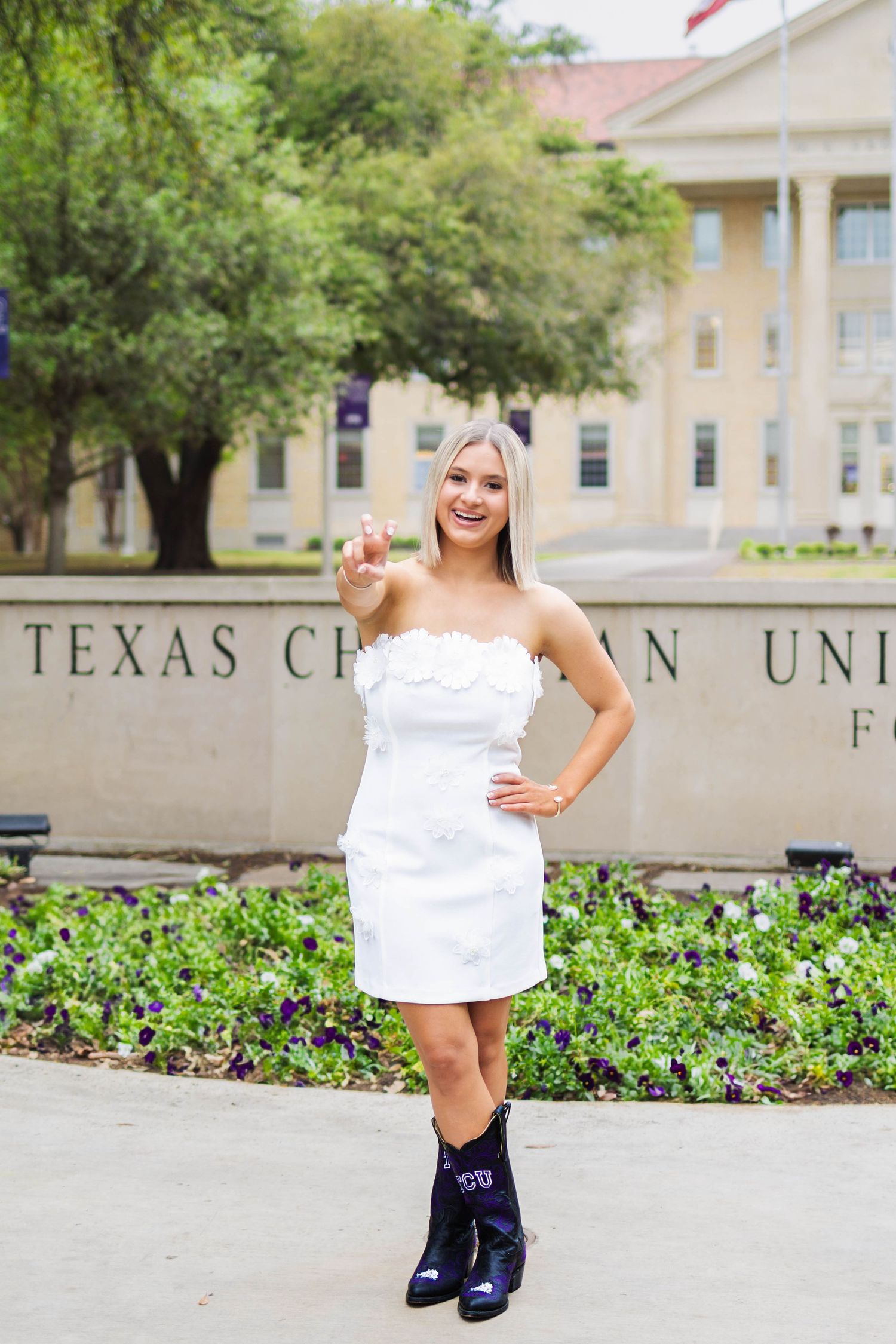 Graduate poses near university signage wearing white dress and purple accessories for graduation photos.