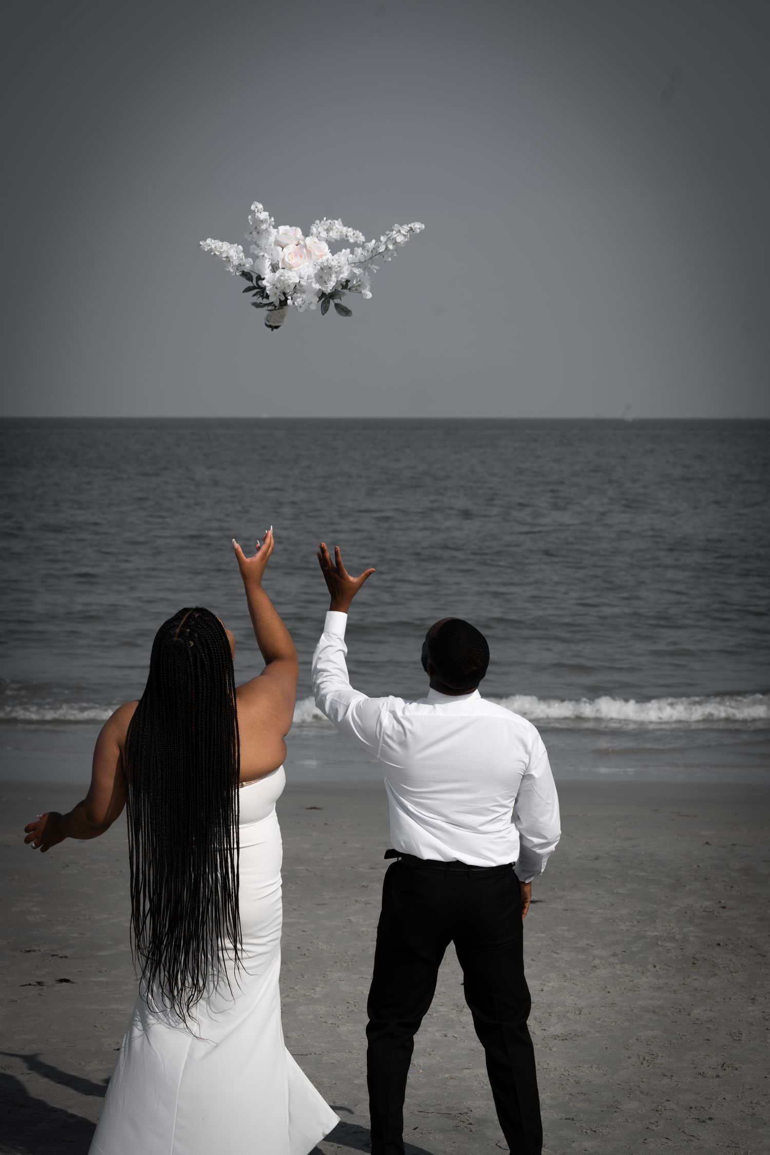 Two people in white shirts stand on a beach watching drones fly over the ocean horizon in a romantic black and white scene.