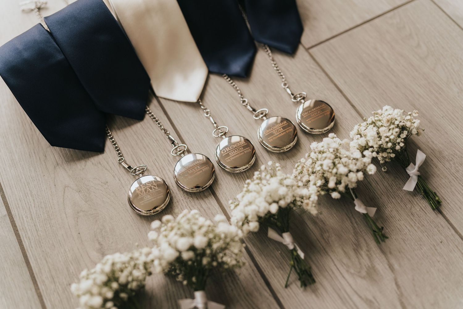 Wedding accessories including white baby's breath flowers, black ties, and pocket watches arranged on a light wooden floor.