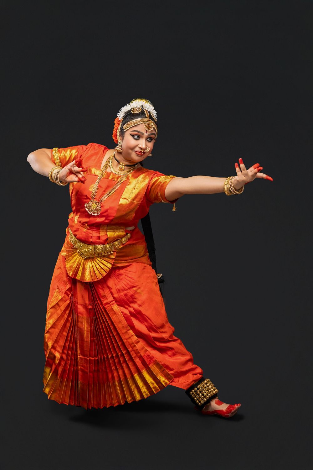 A dancer performs traditional Indian Bharatanatyam dance movements wearing a vibrant orange and gold costume against black backdrop.