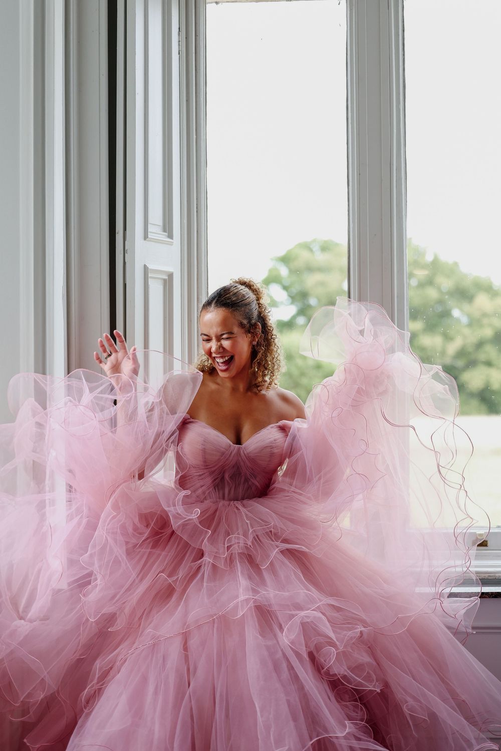 A joyful figure twirls in a voluminous pink tulle gown next to bright windows in an elegant interior setting.